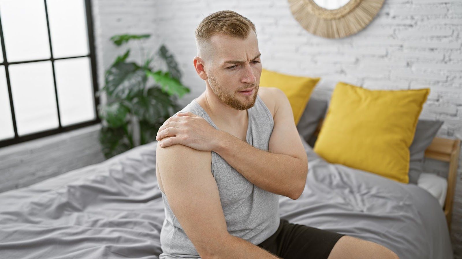 a young man experiences shoulder pain while sitting on a bed